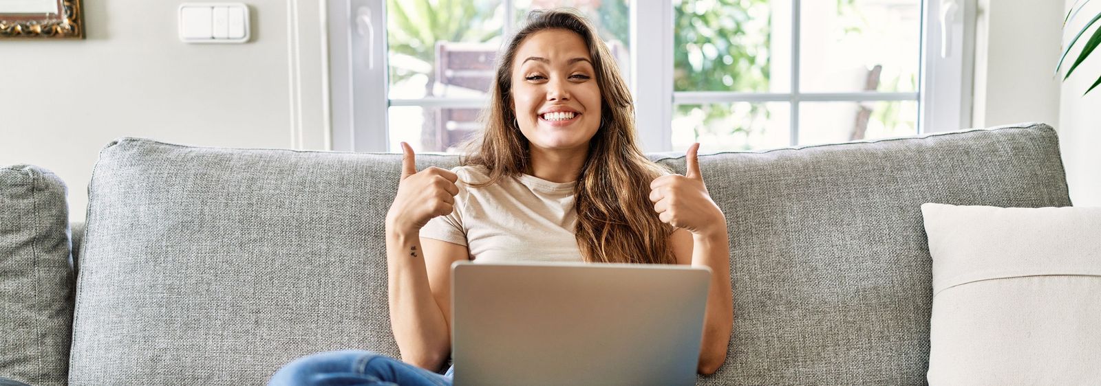 A young woman sits cross-legged on the sofa with a laptop on her lap, smiling at the camera with both thumbs up.