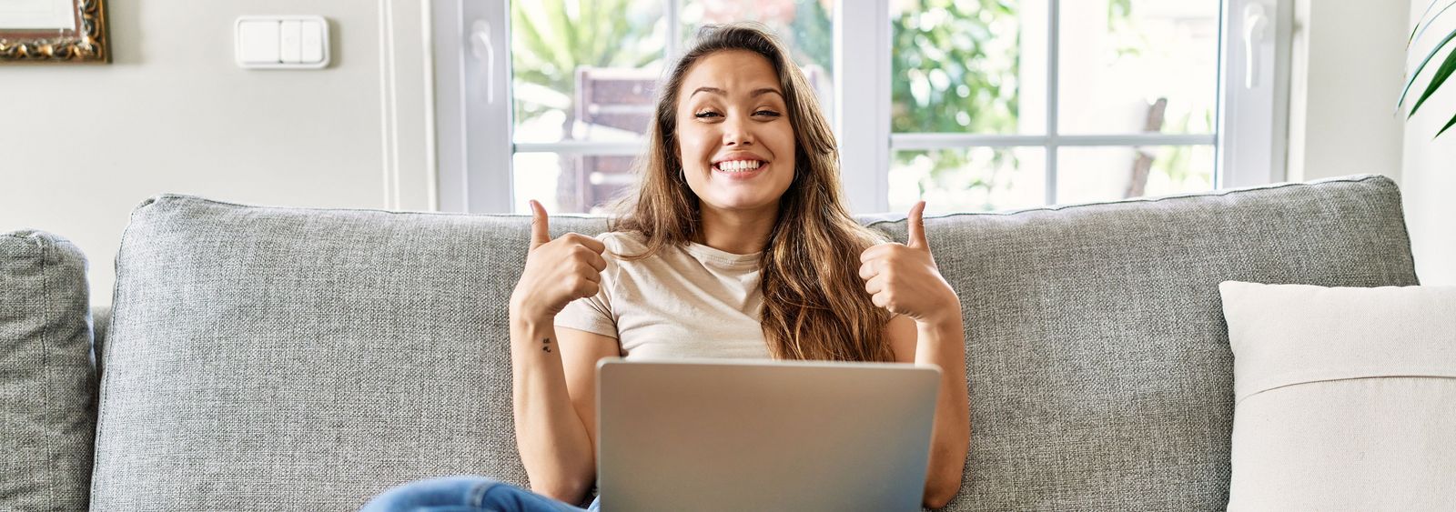 A young woman sits cross-legged on the sofa with a laptop on her lap, smiling at the camera with both thumbs up.