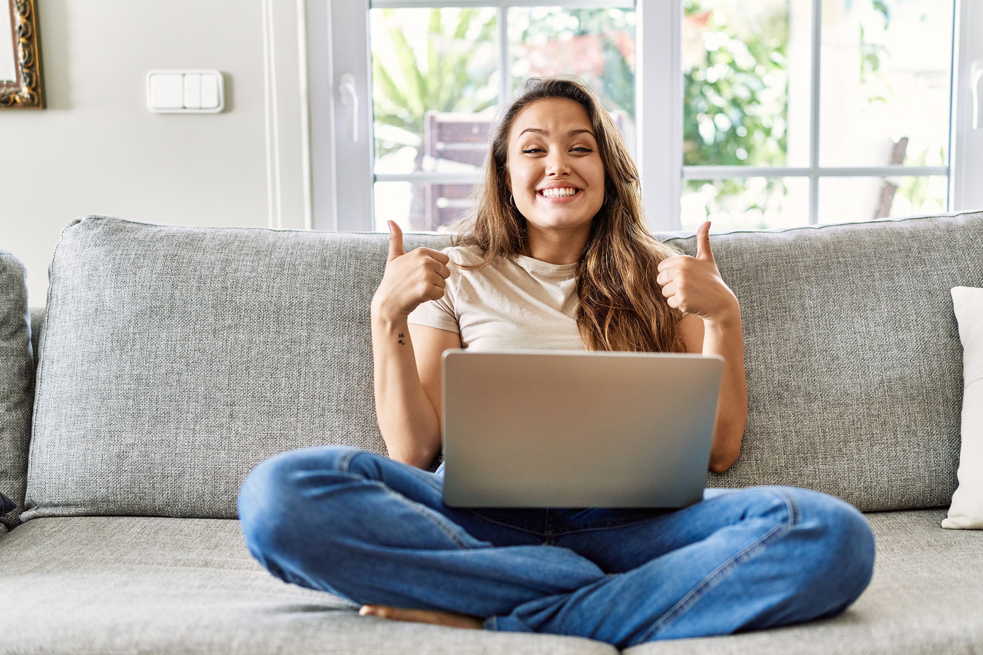 A young woman sits cross-legged on the sofa with a laptop on her lap, smiling at the camera with both thumbs up.